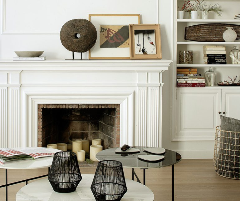 Sala de estar con chimenea blanca decorada con cuadros, velas y estanterías llenas de libros y objetos decorativos.